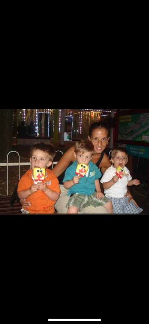 Woman with three young children holding colorful toys indoors with decorative lights in background